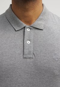 Harmaa poolopaita, jossa on teksturoitu neulos, kaksinappinen aukko ja pieni tunnus vasemman rinnan kohdalla. Kauluksellinen malli.