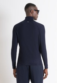 Maglione a collo alto ribbed in navy con maniche lunghe e silhouette aderente, mostrato di spalle, con un colletto alto e un branding discreto.