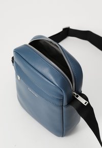 Borsa a tracolla in pelle blu con una superficie liscia, chiusura a zip e tracolla in tessuto nero. Presenta una tasca frontale e un dettaglio di marchio in argento.
