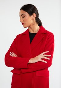 Cappotto rosso su misura con collo a scomparsa, grandi tasche frontali e un tessuto strutturato. Il modello mostra le braccia incrociate, evidenziando il design del cappotto.