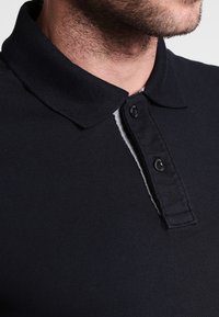 Tmavě modré polo tričko s klasickým límečkem, dvoubarevným lemem a texturovanou tkaninou. Límeček je dobře definovaný s jemným šedým akcentem.