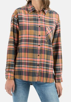 Femme portant une chemise à manches longues à carreaux dans des tons marron, rose, bleu et orange avec une poche poitrine, associée à un jean bleu clair.