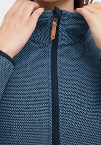 Veste en maille bleue avec un col haut, dotée d'une fermeture éclair et d'un tissu texturé. Accentuée par un tirant de fermeture éclair en cuir marron.