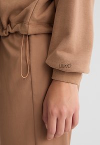 Felpa in cotone marrone con cintura elastica, dotata di un cordoncino e di un piccolo logo "LIU JO" ricamato lungo il polsino della manica.