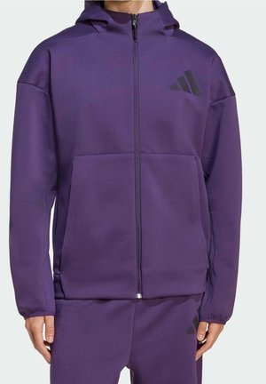 Osoba w fioletowej kurtce z kapturem na suwak i dopasowanych spodniach z czarnym logo Adidas na klatce piersiowej i udzie, stojąca z rękami rozluźnionymi.