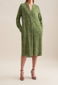 Zelené šaty do délky ke kolenům s výstřihem do V, dlouhými rukávy, bílým puntíkovaným vzorem a bočními kapsami, doplněné černými otevřenými sandály na vysokém podpatku.