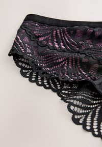 Crne čipkaste gaćice s zamršenim cvjetnim uzorcima i nazubljenim rubovima, na kojima se ispod vidi ružičasta tkanina, ravno položene na neutralnoj površini.