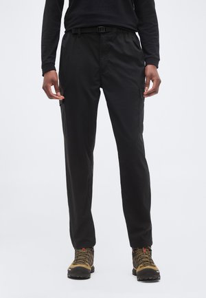 Persoană purtând pantaloni negri cargo cu buzunare laterale, o cămașă neagră cu mâneci lungi și bocanci maro de drumeție, stând pe un fundal simplu.