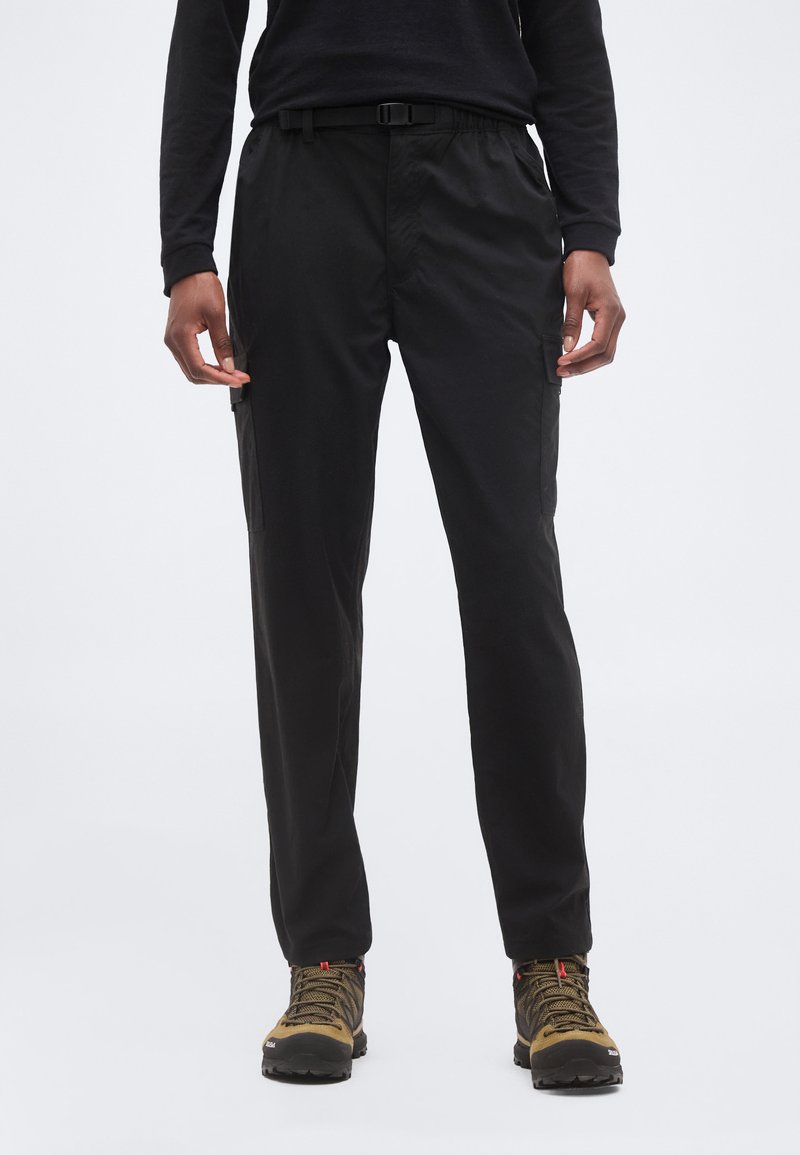 Άτομο που φοράει μαύρο παντελόνι cargo με πλαϊνές τσέπες, μαύρο μακρυμάνικο πουκάμισο και καφέ πεζοπορικά μποτάκια, στέκεται σε απλό φόντο.