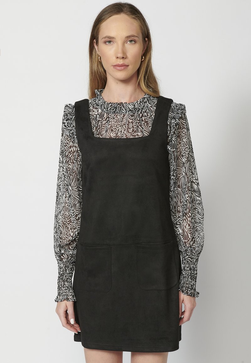 Robe en daim noir avec un décolleté carré, dotée de deux poches avant. En dessous, un chemisier transparent noir et blanc à motifs avec des manches à volants.