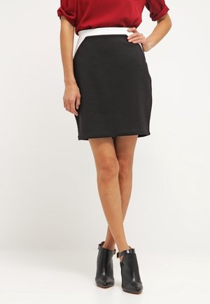 Jupe mini noire texturée avec une ceinture blanche et des accents sur les côtés, associée à des bottines noires. Blouse rouge avec des manches retroussées.
