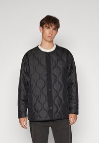 Black quilted jacket with a round neckline and snap button closure. Features a textured pattern and long sleeves. Worn over a white shirt.