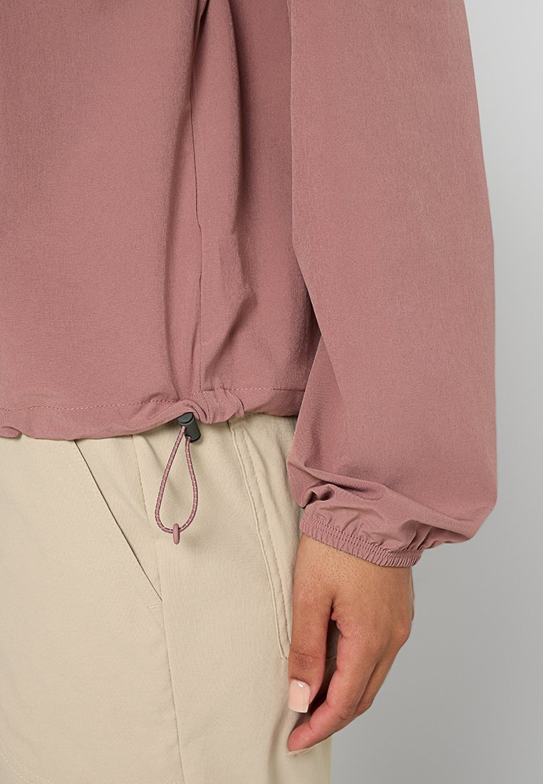 Długo rękawowa, skrócona bluzka z miękkiego w kolorze mauve materiału, z elastycznymi mankietami i regulowanym sznurkiem u dołu dla lepszego dopasowania. Połączona z beżowymi spodniami.