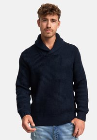 Granatowy sweter wykonany na drutach z kołnierzem szalowym, z teksturowanym wzorem oraz ściągaczami na rękawach i u dołu. Noszony z jasnoniebieskimi, postarzanymi dżinsami.