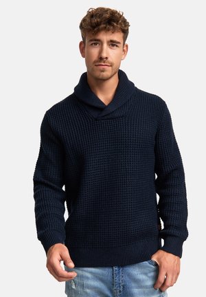 Suéter de punto azul marino con cuello de chal, que presenta un patrón texturizado y puños y dobladillo acanalados. Llevado con jeans desgastados de color azul claro.