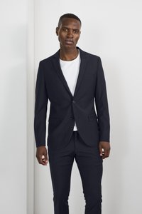 Un costume bleu marine avec une coupe slim, une veste à un bouton et un pantalon assorti. En dessous, un t-shirt blanc à col rond est porté.