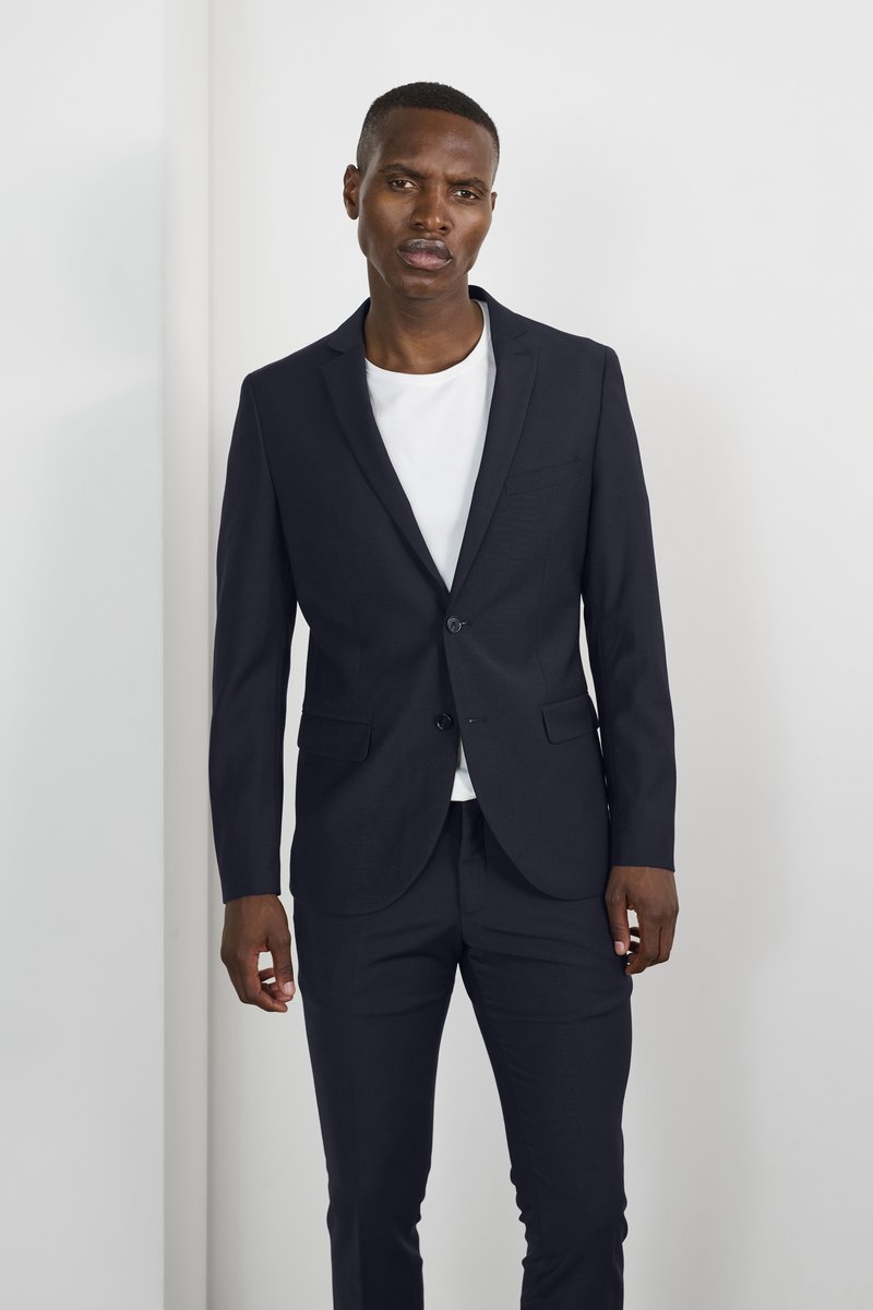 Un costume bleu marine avec une coupe slim, une veste à un bouton et un pantalon assorti. En dessous, un t-shirt blanc à col rond est porté.