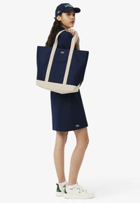 Sac fourre-tout en toile bleu marine avec des anses beiges, arborant un petit logo vert. Le modèle porte une robe assortie bleu marine et des baskets blanches avec des touches de vert.