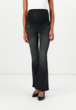 Pregnant person wearing black maternity jeans with an elastic belly panel and black heels, standing against a plain white background.
