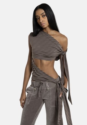 Hosszú fekete hajú nő egyrészes barna csíkos felsőt visel, amelyet deréknál és oldalt megkötött, szürke zsinóros nadrággal párosítva.