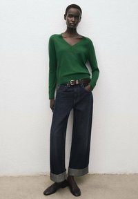 Maglione verde a scollo a V, pantaloni di jeans a gamba larga e orli risvoltati di colore scuro, cintura di pelle marrone e scarpe marroni. Texture lisce e forma aderente.