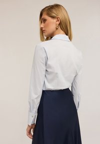 Camicia a righe azzurro chiaro con colletto classico, maniche lunghe e polsini con bottoni, abbinata a una gonna nera scura, texture liscia.