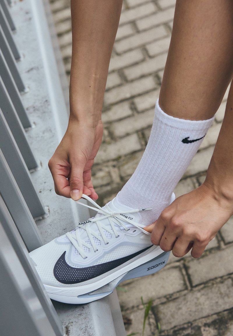 Bijela Nike Zoom Fly trkačka cipela s sivim detaljima, vezana čipka, nosi se s bijelim rebrastim čarapama, ruka podešava cipelu na asfaltiranoj površini.