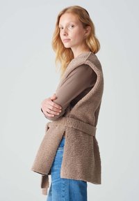 Beige Fleece-Weste mit offenen Seiten und einem Bindegürtel in der Taille, getragen über einem braunen Langarmoberteil, kombiniert mit blauen Jeans.