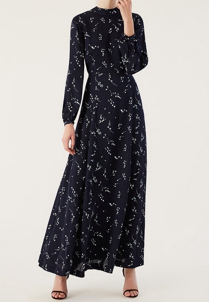 Femme portant une robe longue à manches longues bleu marine avec un petit motif floral blanc et des talons ouverts noirs, debout avec une main près du visage.