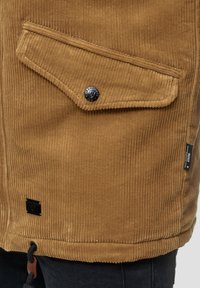 Bolso de chaqueta de pana marrón con solapa y botón a presión negro, etiqueta negra en el lado y dobladillo con cordón sobre pantalones oscuros.
