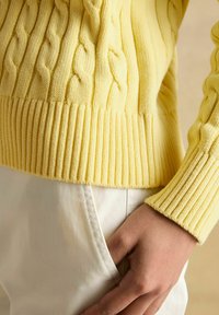 Artimas plano kadras rodo ranką, laikomą baltoje kelnėje, vilkint geltoną megztinį su kabliukais, turiu gūžteles ir apykakle.