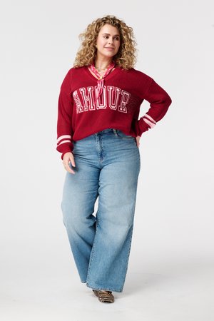 Femme aux cheveux bouclés portant un pull rouge avec l'inscription "AMOUR", une écharpe rose, un jean large et des chaussures à imprimé zèbre, debout la main sur la hanche.