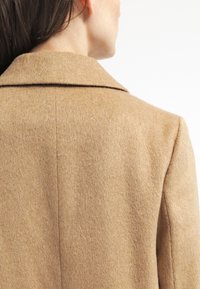 Světlý hnědý vlněný kabát s klasickým límcem, hladkou texturou a jednoduchým střihem, s viditelnými švy na zadní straně.