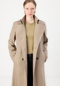 Beige trench coat with a double-breasted design, button accents, and side pockets, worn over a light green top and dark jeans.