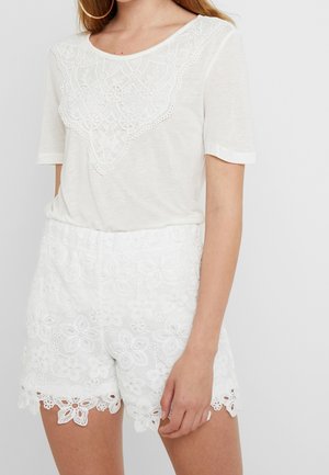 Frau trägt ein weißes Kurzarmoberteil mit Spitzenpasse und hoch taillierte weiße Shorts aus floraler Spitze.