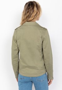 Giacca in cotone verde oliva con vestibilità su misura, caratterizzata da pattine sulle spalle e una texture liscia. La vista posteriore mostra un orlo curvo.