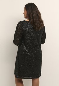 Robe noire à sequins avec un tissu doux et texturé, des manches courtes et une coupe décontractée, se terminant au-dessus du genou. La surface scintillante reflète la lumière.