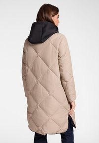 Béžový prošívaný kabát s diagonálním prošíváním a černou kapucí. Kabát má dlouhý střih a boční rozparky, je vyroben z hladké tkaniny.