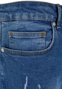 Jean en denim bleu avec une poche arrière, présentant des coutures visibles et des détails de déchirure. Le tissu affiche une texture délavée.