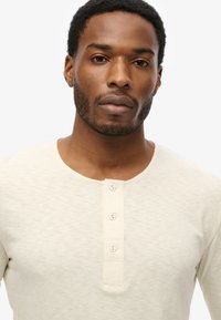 Camisa henley de manga larga en un tono beige claro, con un cuello redondeado y una tapeta con tres botones. Tejido suave y texturizado con un ligero efecto jaspeado.