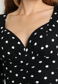 Czarna bluzka w białe kropki, z głębokim dekoltem w kształcie V, marszczoną strukturą i dopasowanym krojem, podkreślająca smukłą sylwetkę.