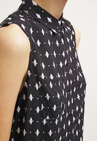 Czarna koszulka bez rękawów z kołnierzykiem, z białym geometrycznym wzorem i małą kieszenią po lewej stronie. Miękka, teksturowana tkanina.