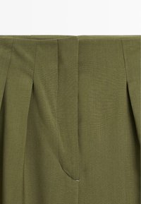 Pantalons ajustés vert olive avec plis sur les côtés, en tissu lisse et texturé, avec des détails de couture discrets le long de la taille.