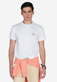 Camiseta blanca de algodón de manga corta, con un pequeño diseño gráfico en el lado izquierdo del pecho. Combinada con un sudadera naranja atada a la cintura.