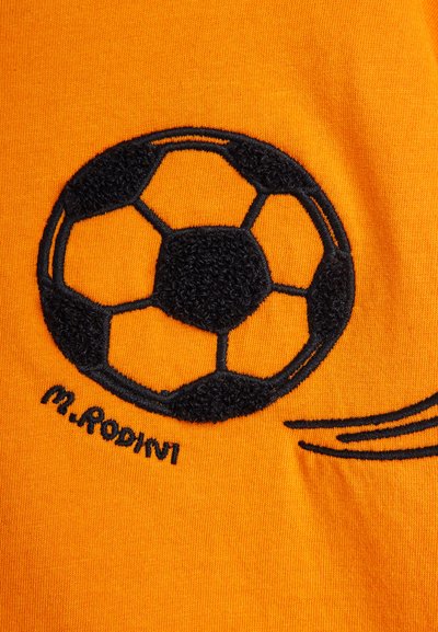 Μαύρη κεντημένη μπάλα ποδοσφαίρου σε πορτοκαλί ύφασμα με μαύρες ραμμένες γραμμές κίνησης και το κείμενο «M. Rodini» κάτω από τη μπάλα.