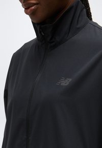 Κοντινό πλάνο προσώπου ατόμου που φοράει μαύρο μπουφάν με φερμουάρ και το λογότυπο της New Balance στο στήθος και ψηλό γιακά, στραμμένο ελαφρώς προς τα αριστερά.