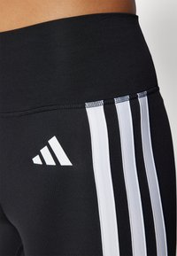 Melnas legingi ar plašu jostasvietu, trīs vertikālām baltām svītrām sānos un baltu Adidas logo. Gluds, elastīgs materiāls.