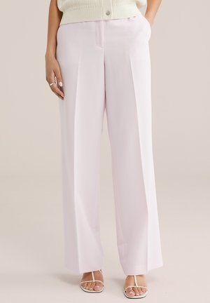 Žena nosi bledo ružové nohavice s vysokým pásom a širokými nohavicami s prednými záhybmi, doplnené bielymi sandálmi na remienky a krémovým pleteným topom.