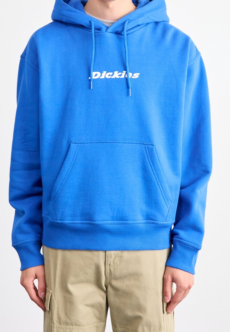 Homme portant un sweat à capuche bleu vif de la marque Dickies avec poche avant et pantalon cargo beige sur fond blanc uni.