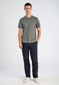 Graues kurzärmliges Poloshirt mit Knopfleiste, strukturierter Stoff, kombiniert mit marineblauen Hosen und weißen Sneakern, steht vor einem schlichten Hintergrund.